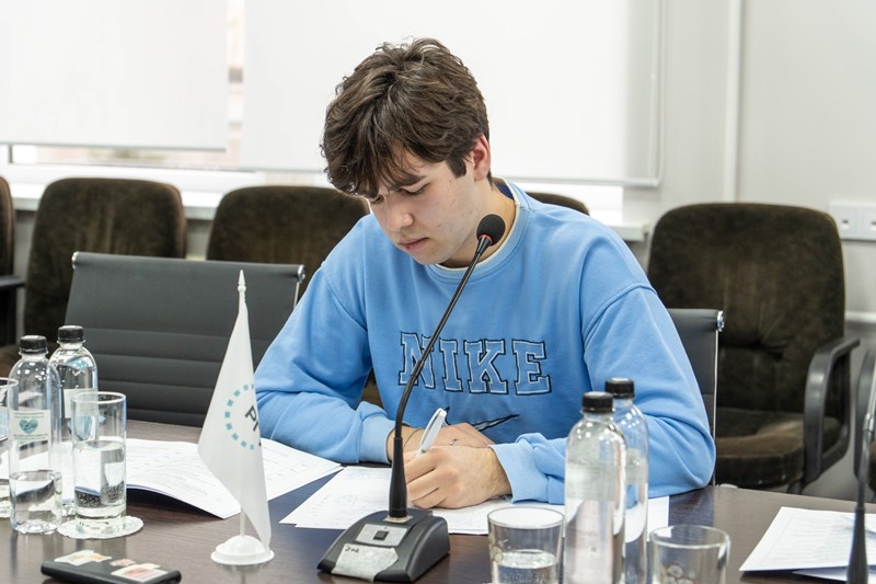 A young man in a blue Nike sweatshirt writing at a conference table with a microphone and documents