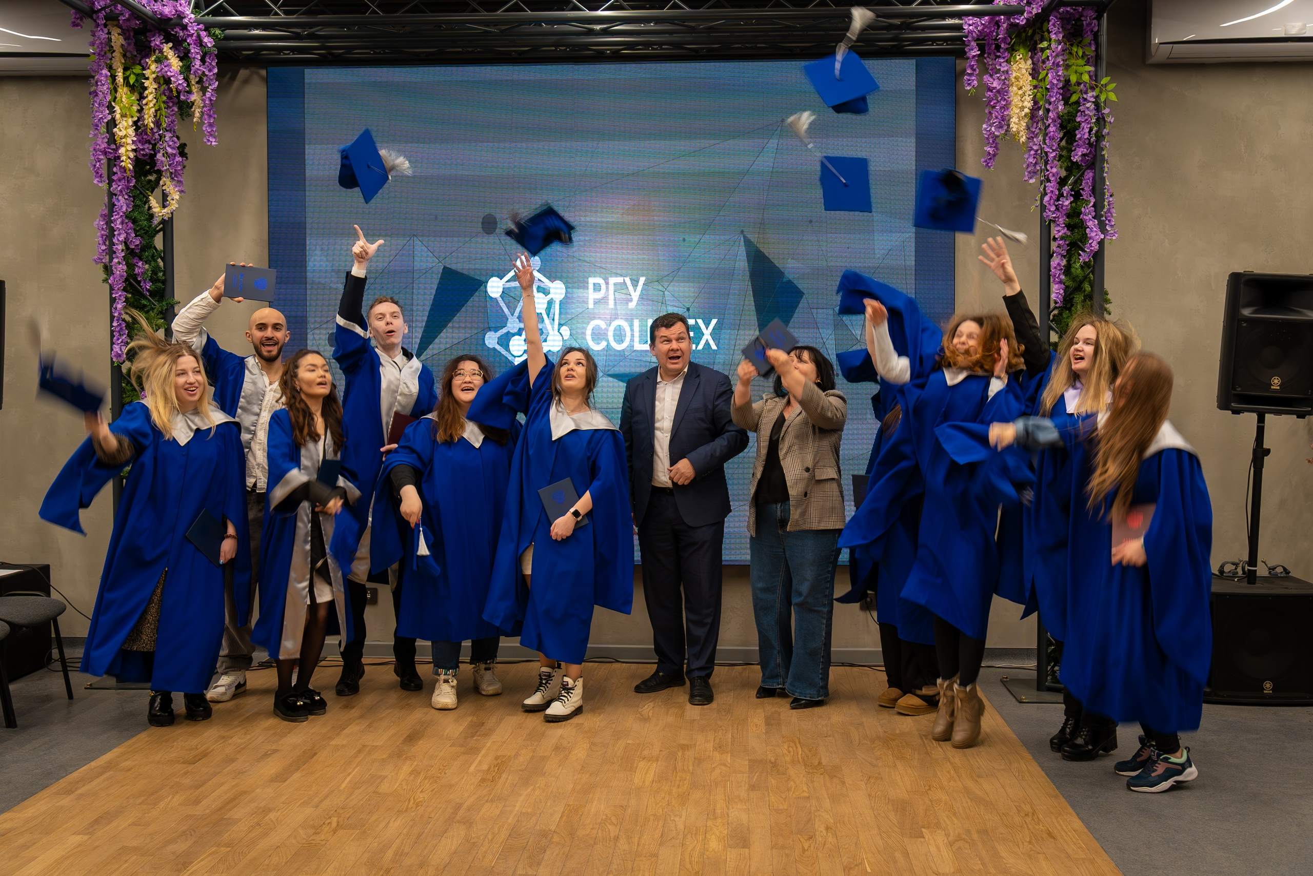 Группа выпускников в синих мантиях радостно подбрасывает шапочки в воздух на сцене на фоне экрана с надписью РГУ СоцТех