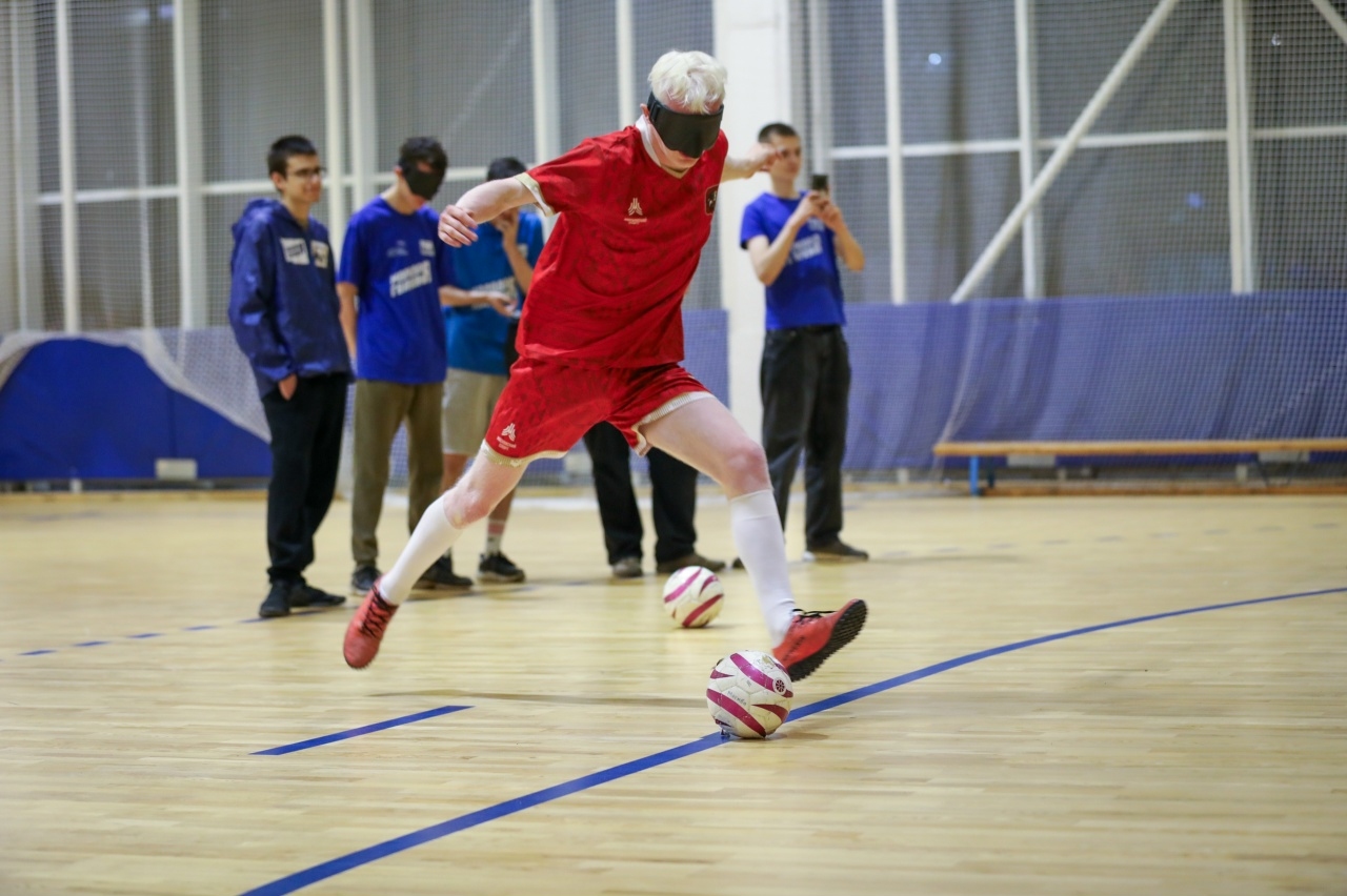 Спортсмен с повязкой на глазах играет в футбол в спортивном зале, а другие участники наблюдают за игрой