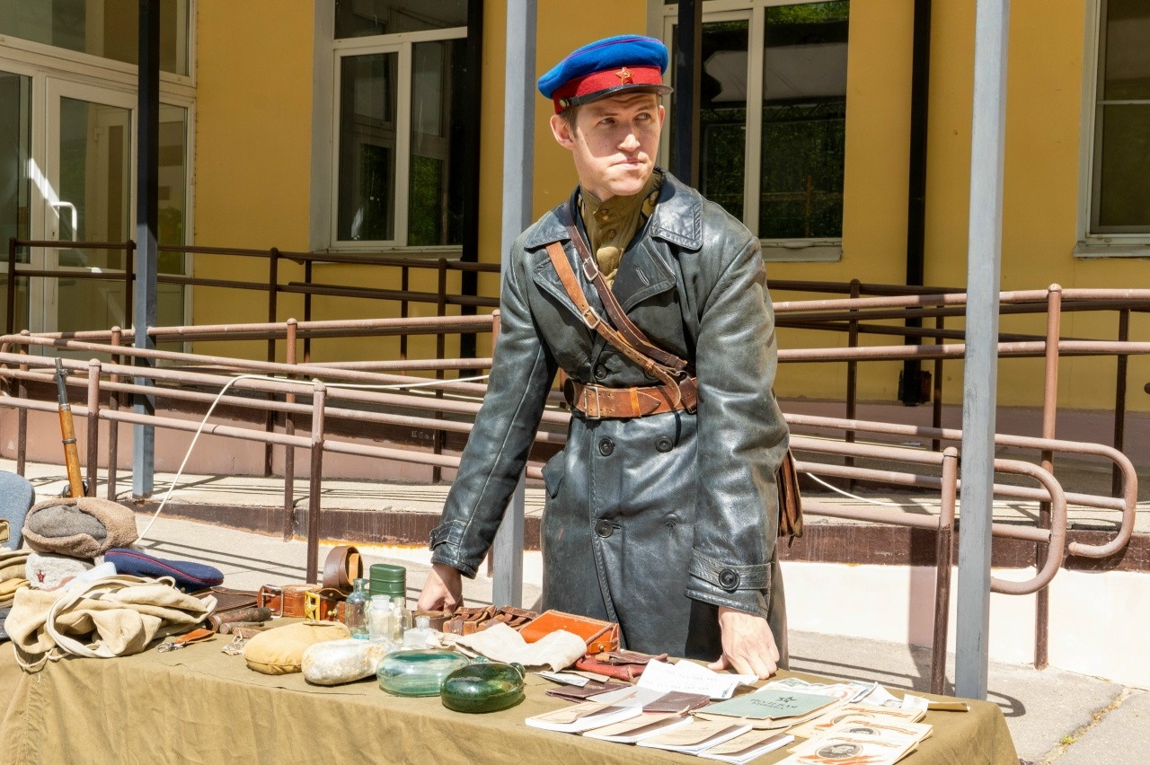 Молодой человек в форме советского командира (кожаная шинель и будёновка с красной звездой) стоит за столом с военными артефактами и документами