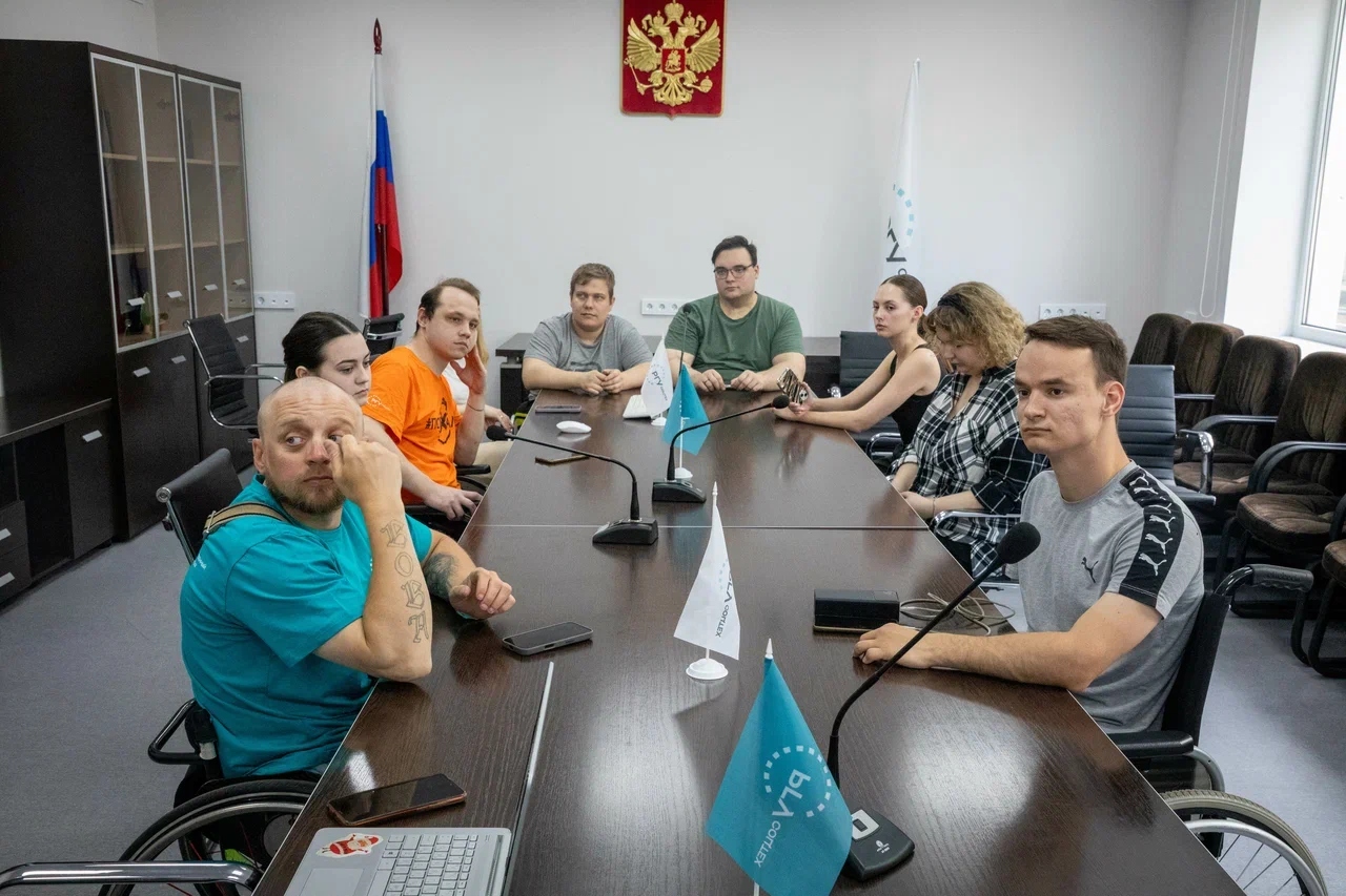Группа из восьми человек сидит за длинным столом в конференц-зале с гербом и флагом России на стене; двое мужчин на переднем плане находятся в инвалидных колясках, на столе стоят микрофоны и небольшие флажки