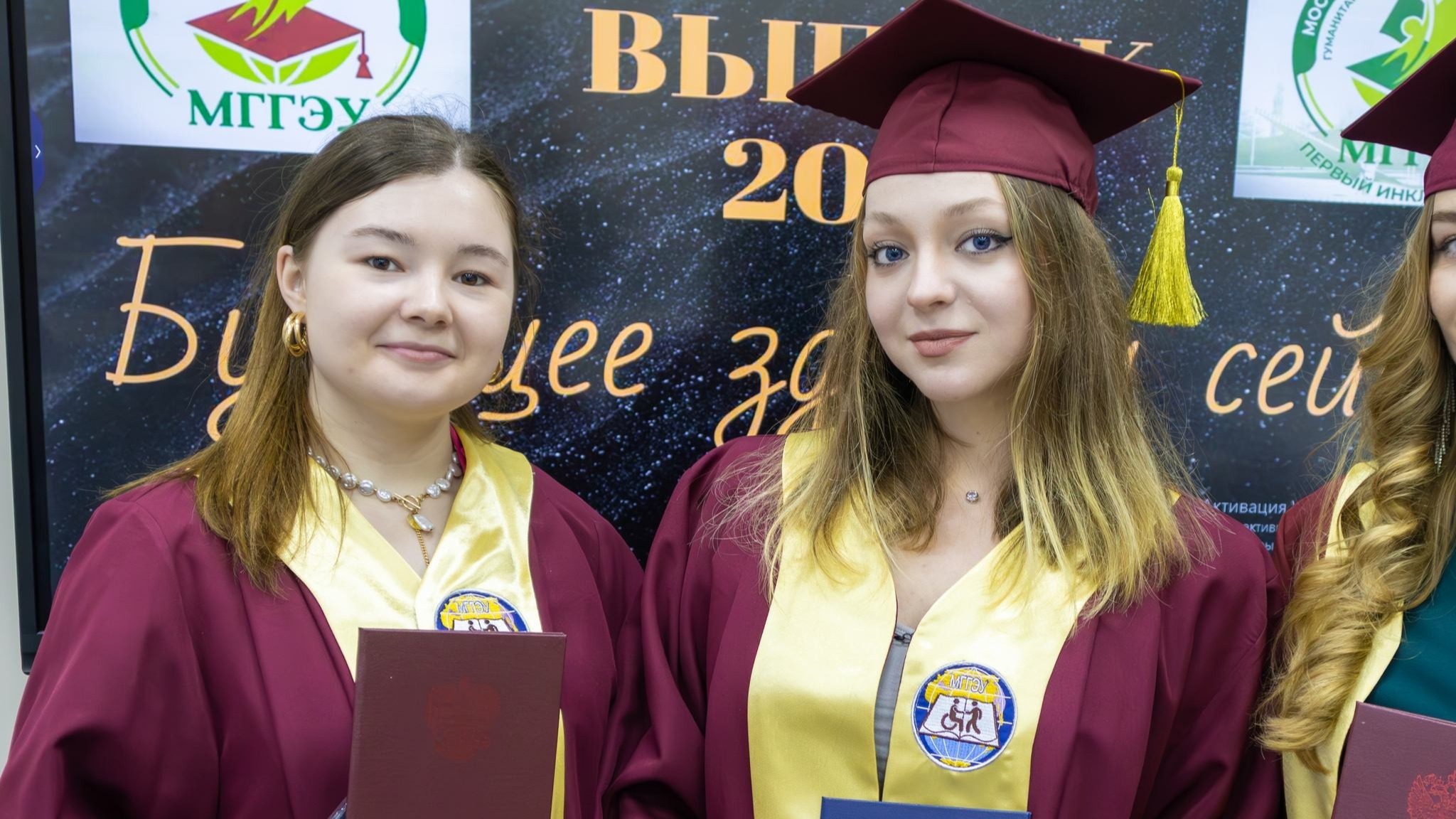 Две выпускницы в бордовых мантиях держат дипломы и улыбаются на фоне праздничного баннера