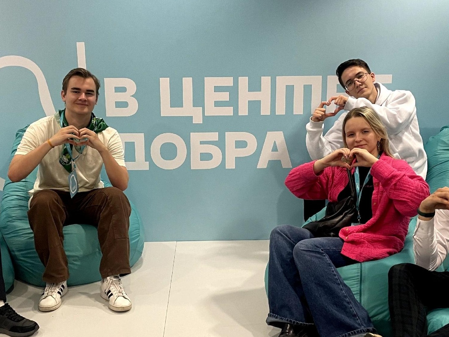 Трое молодых людей сидят на бирюзовых пуфах перед стеной с надписью «В ЦЕНТРЕ ДОБРА» и показывают жест «сердце» руками