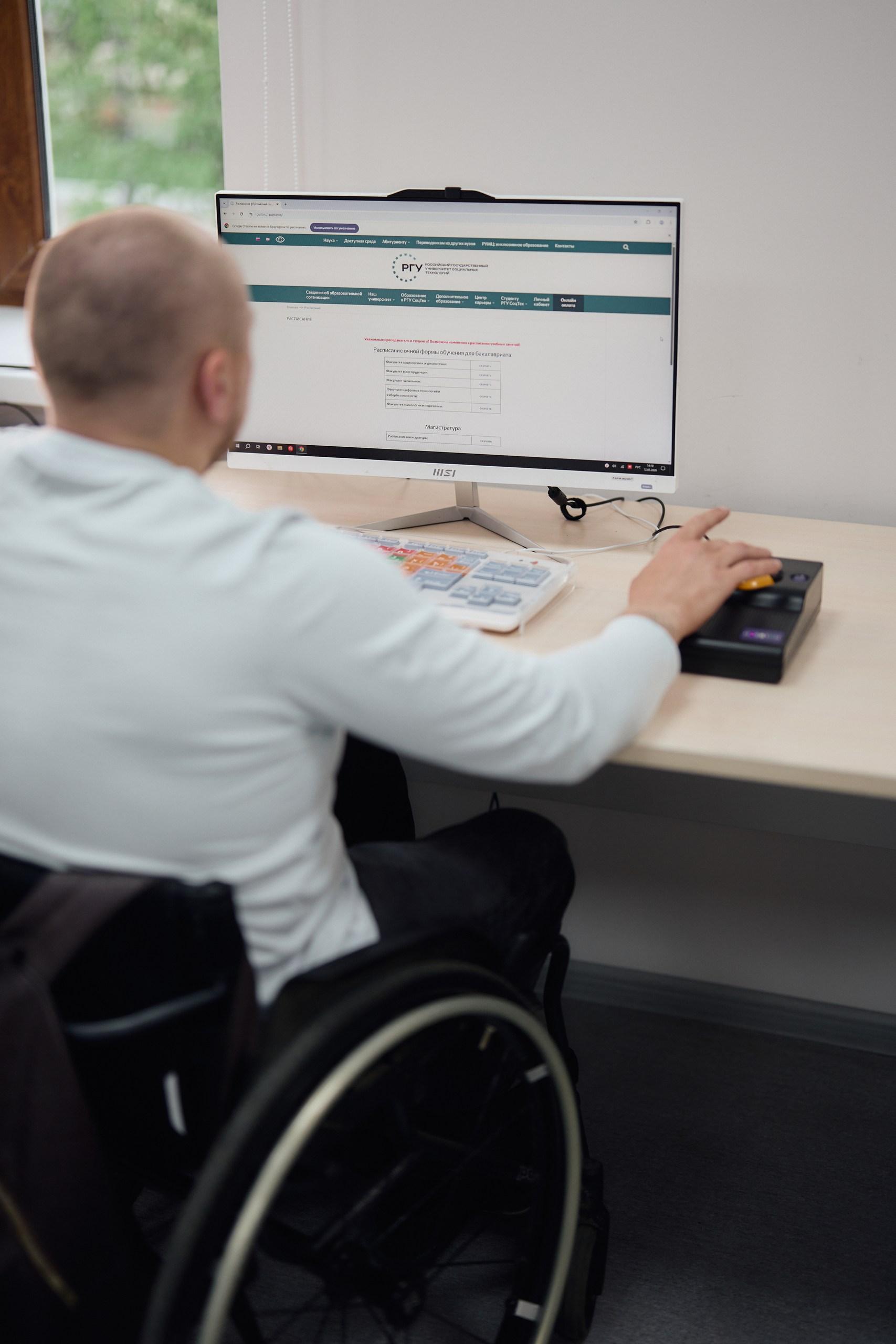 Un homme en fauteuil roulant utilise un clavier et un trackball spécialisés pour naviguer sur le site web de l'Université d'État russe des technologies sociales sur un ordinateur de bureau.