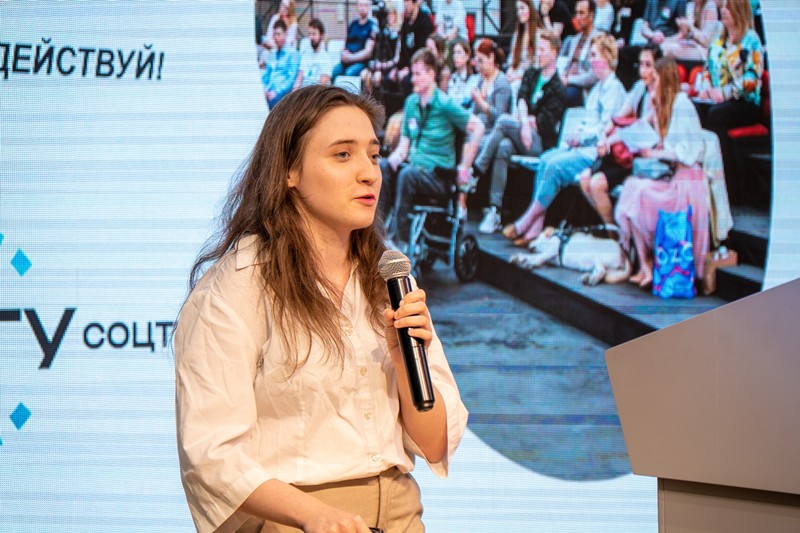 A girl in a white shirt is speaking with a microphone on stage in front of a screen with the RSU SOCTECH logo and the inscription 'DEYSTVUY!'