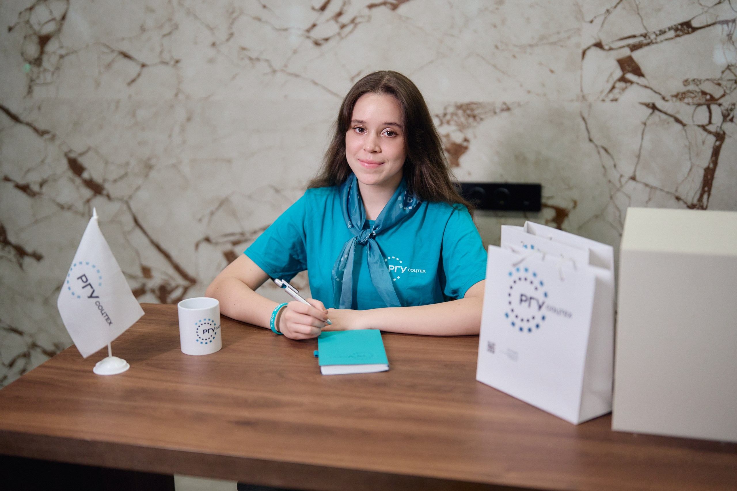 Девушка в бирюзовой футболке с логотипом РГУ СОЦТЕХ сидит за столом с брендированными сувенирами и блокнотом