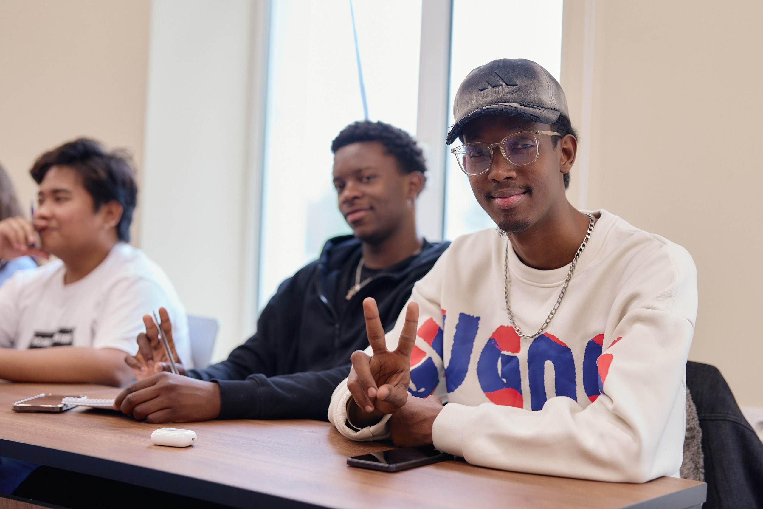Trois jeunes hommes assis à une table dans une salle de classe ; l'étudiant au premier plan portant une casquette et des lunettes sourit et fait le signe de la paix vers l'appareil photo