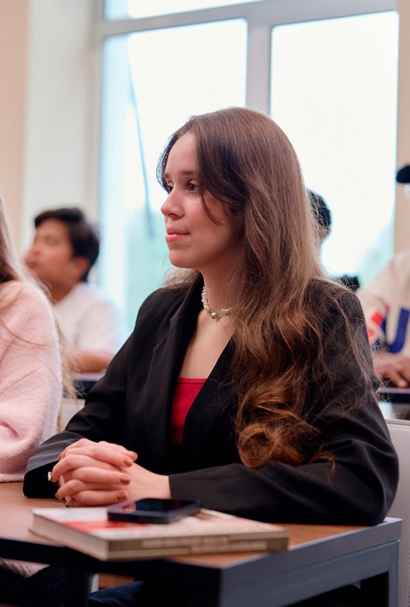 На изображении Молодая девушка сидит за партой в аудитории и слушает преподавателя, перед ней лежат книги и смартфон.
