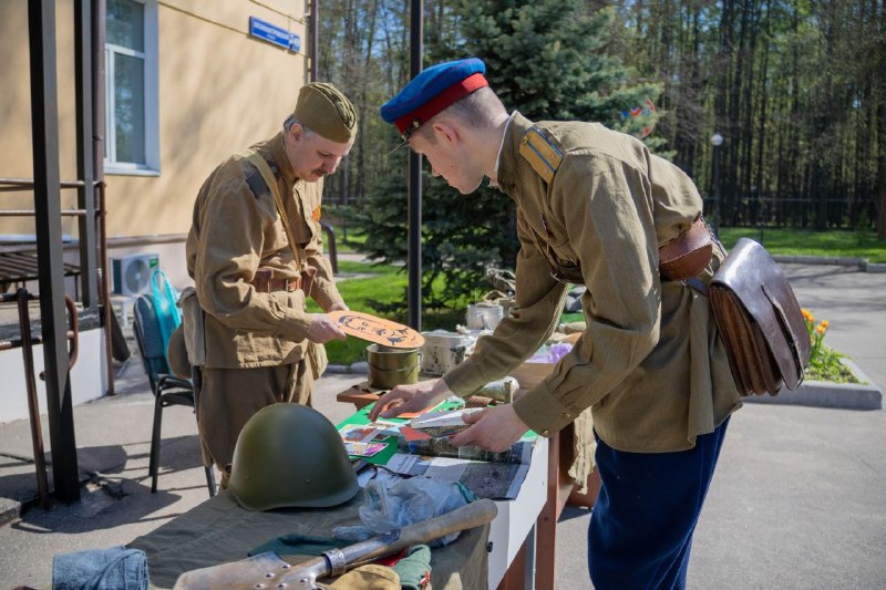 Два человека в советской военной форме осматривают исторические экспонаты, включая каску и документы, на уличном столе