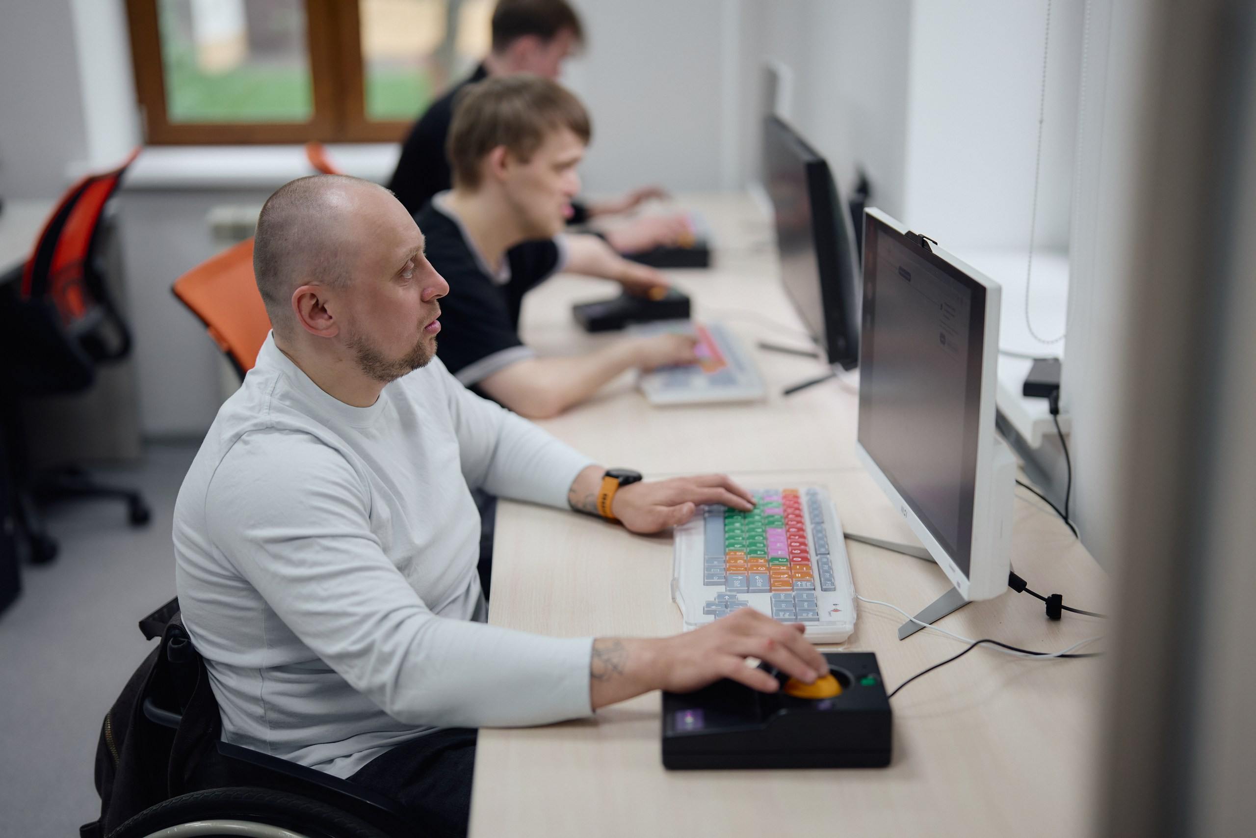 Мужчина в инвалидной коляске работает за компьютером, используя специальную цветную клавиатуру и трекбол