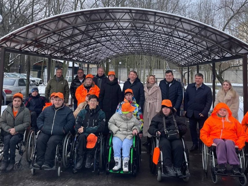 Групповое фото людей в инвалидных колясках и стоящих рядом организаторов под навесом на улице, многие одеты в оранжевые куртки и кепки