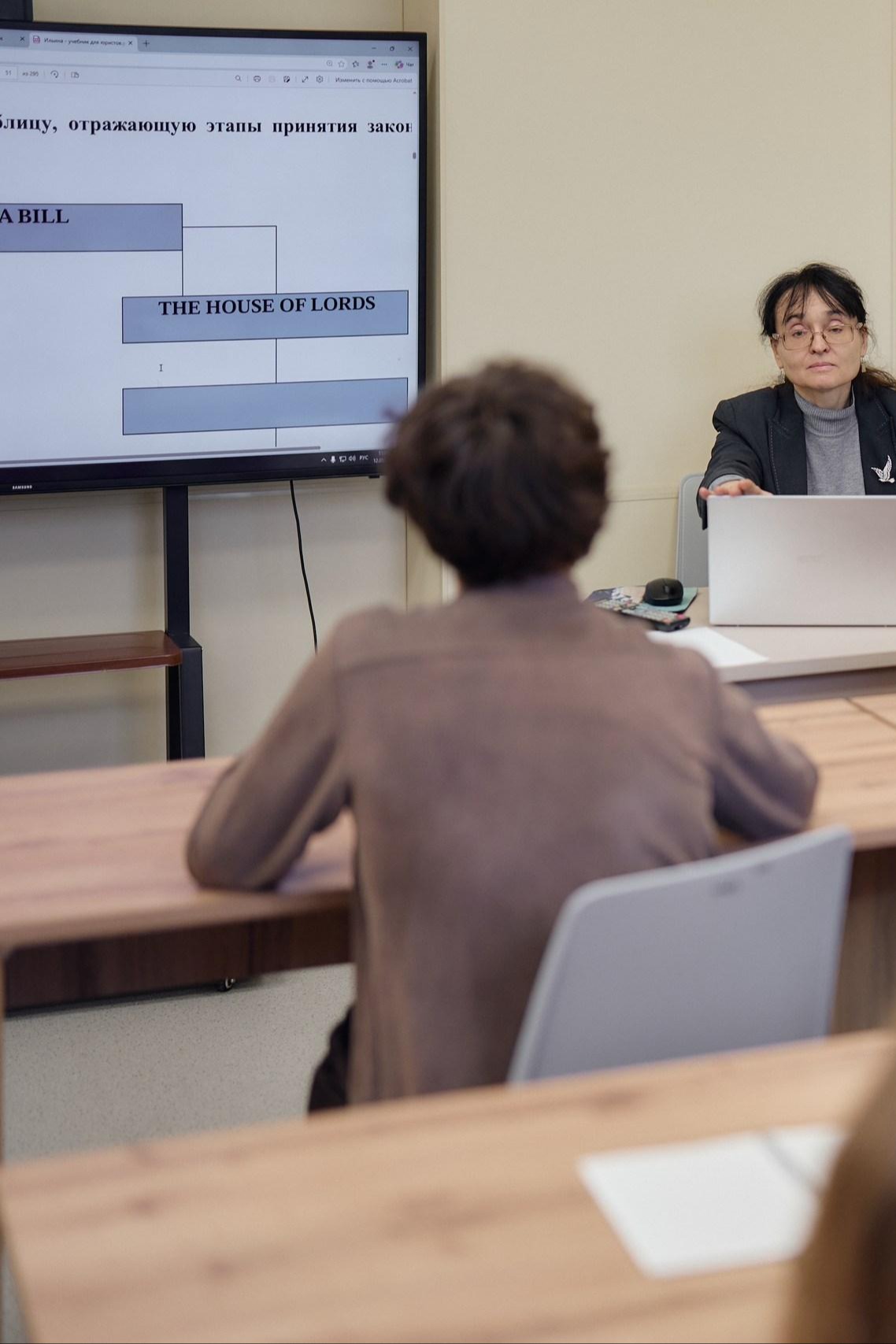 Преподавательница проводит лекцию в аудитории, позади нее презентационный экран