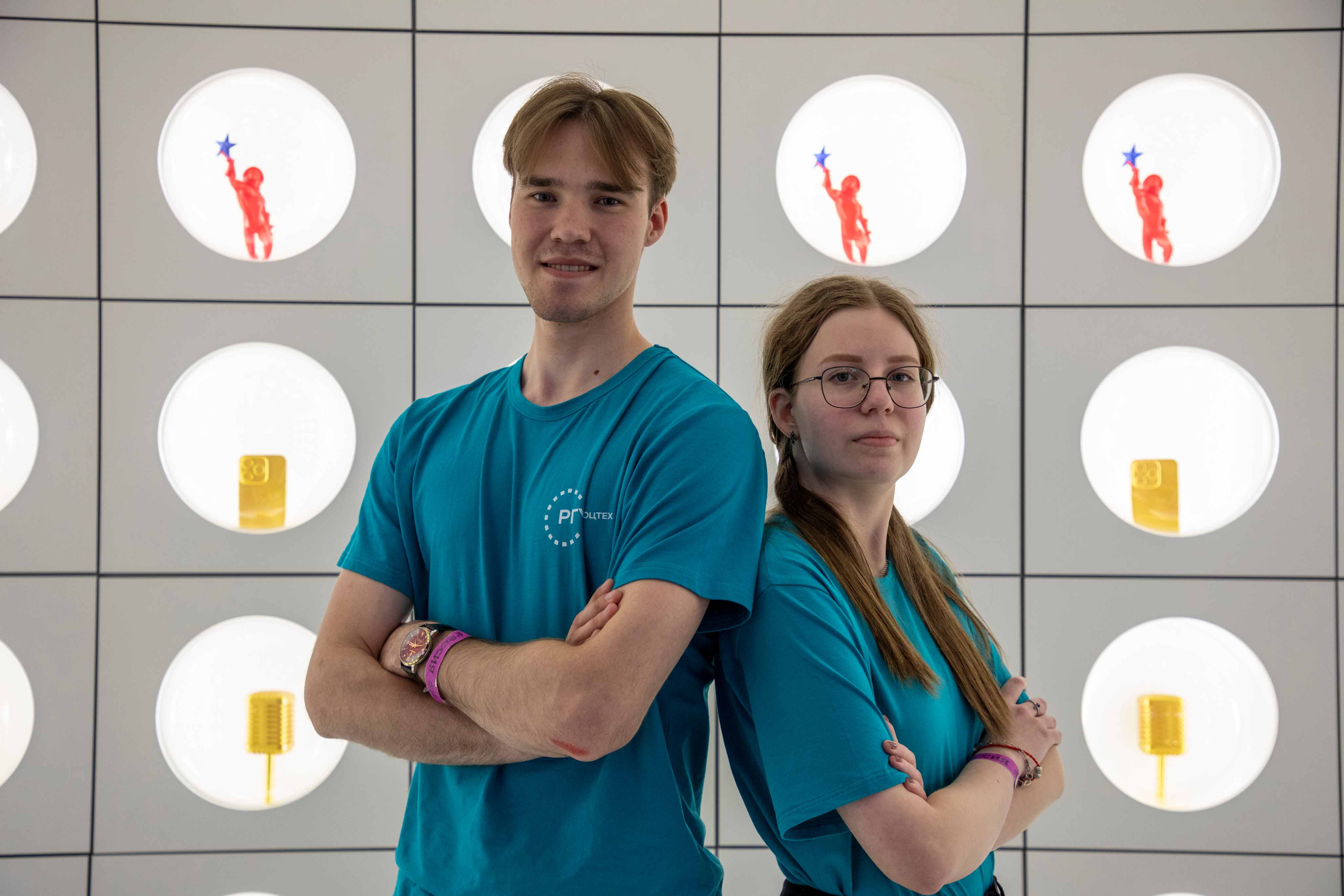 Two students wearing matching turquoise RSU ST t-shirts posing in front of a modern illuminated wall.