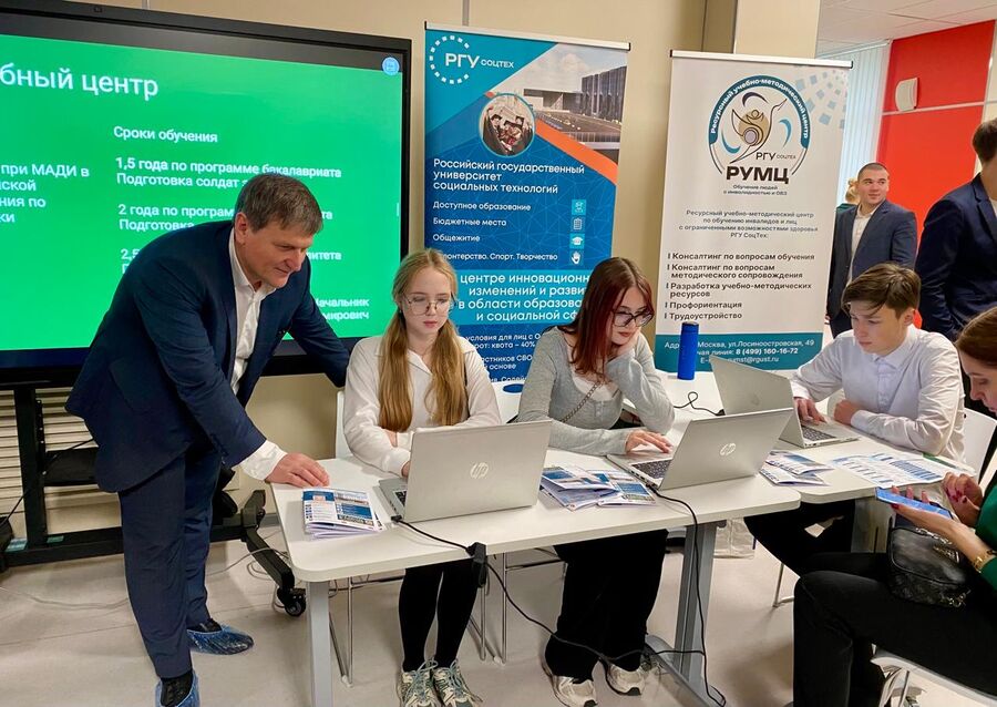 A man in a suit is consulting students at a table with laptops, against the background of RSU SOC TECH banners and a screen with information about education.